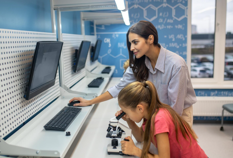 A woman stands at a computer desk while a girl in a pink shirt looks through a microscope in a science classroom.